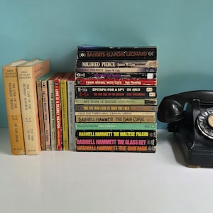Puede incluir: Una pila de libros de bolsillo vintage variados con lomos de diversos colores y títulos que incluyen Bandits, Mildred Pierce y The Maltese Falcon, junto a un teléfono de disco negro con un auricular descansando sobre la parte superior.