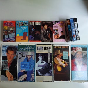 May include: A collection of VHS tapes featuring country music artists, including Garth Brooks, Randy Travis, George Strait, and Alan Jackson. The tapes are arranged in a row on a light brown surface.