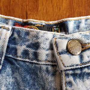 May include: Close-up of the waistband of a pair of light blue denim jeans with a gold button that reads "Gasson New York".