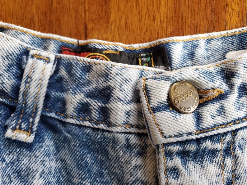 May include: Close-up of the waistband of a pair of light blue denim jeans with a gold button that reads "Gasson New York".