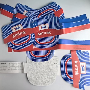 May include: A collection of Amtrak-branded paper items. The designs feature blue and white striped patterns, red accents, and the Amtrak logo. Some pieces include the text "Amtrak" and "All Aboard... If You Can Find 'Em!"