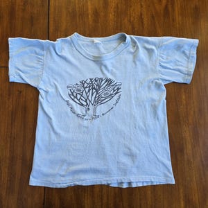 Puede incluir: Camiseta azul claro con cuello redondo y mangas cortas. La parte delantera presenta un gráfico negro de un árbol con bocadillos de texto y la inscripción "Bryn Mawr College 1980 Summer Institute". La tela es suave y desgastada.