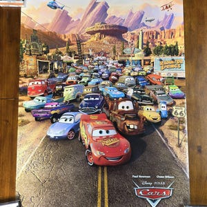 May include: A colorful poster for the Disney Pixar movie "Cars" featuring a large group of anthropomorphic cars on a desert road. The poster includes the text "Radiator Springs" and "Route 66". The movie title and release date are also visible.