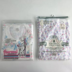 Puede incluir: Dos paquetes de ropa interior térmica. Un paquete contiene una camiseta blanca de manga larga con un estampado floral y el texto "LADIES LONG SLEEVE SHIRT". El otro paquete contiene un pantalón hasta el tobillo con un estampado floral.