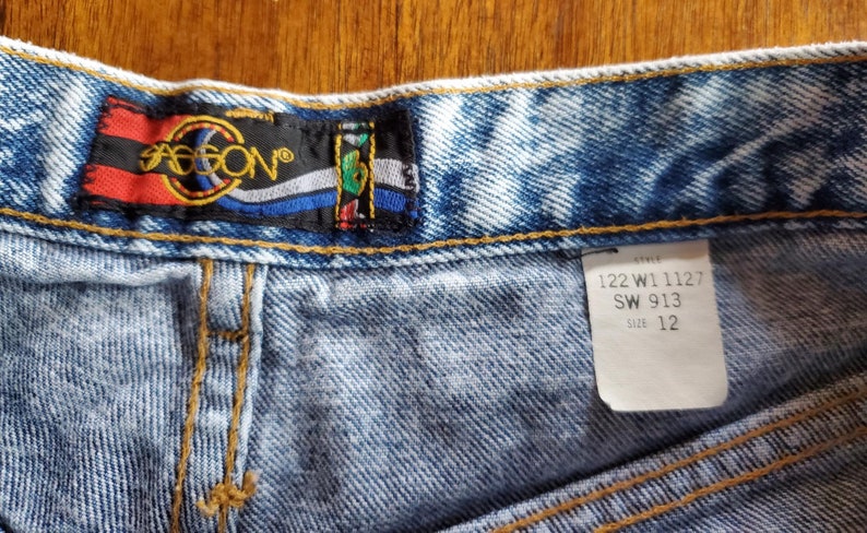 May include: Light blue denim shorts with a faded look. The shorts have a brown leather patch with the word "JASON" in white lettering. The size tag reads "122 W1 1127 SW 913 Size 12".