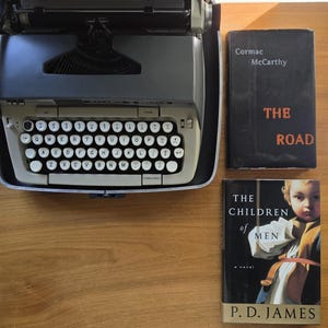 May include: A vintage gray typewriter with white keys and a black ribbon, next to two books. One book is titled "The Road" by Cormac McCarthy, and the other is "The Children of Men" by P.D. James.