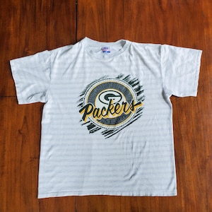 May include: A white striped t-shirt with a green and yellow Green Bay Packers logo. The logo says "National Football League" and "Packers Est. 1919".