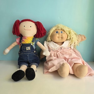 Puede incluir: Dos muñecas vintage. Una muñeca tiene pelo de hilo rojo, una camiseta teñida con tinte, un peto vaquero y zapatos negros. La otra muñeca tiene pelo de hilo rubio y un vestido rosa. Ambas muñecas están sentadas.