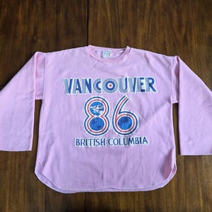 May include: A light pink long-sleeved shirt with the text "VANCOUVER 86 BRITISH COLUMBIA" in blue and white. The number "86" is in a blue and white design. The shirt is made of a soft material.