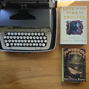 May include: A vintage SCM Galaxie typewriter with white keys and a black frame sits on a wooden surface, next to two books. The books are titled "Love in the Time of Cholera" and "The General in His Labyrinth" by Gabriel García Márquez.