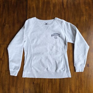 May include: Light grey Champion sweatshirt with the word "MARQUETTE" in navy blue across the chest. The sweatshirt has a crew neck and long sleeves. The university logo is below the word "MARQUETTE".