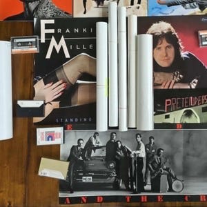 May include: A collection of vintage music memorabilia, including vinyl records, cassette tapes, and rolled posters. The album covers feature various artists and album titles. The items are arranged on a wooden surface.