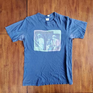 Puede incluir: Una camiseta azul con una imagen descolorida de tres personas en una banda. La imagen está en un rectángulo con el texto "The Allman Brothers Band" encima.