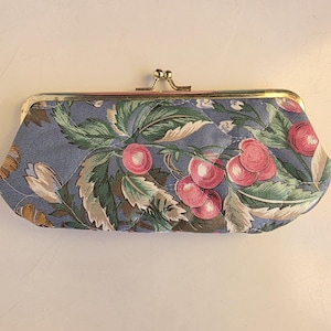 May include: A blue clutch with a gold clasp, featuring a floral pattern of cherries and green leaves. The fabric has a quilted texture. The clutch is a small, handheld accessory.