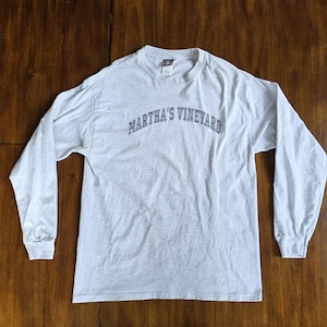 Könnte beinhalten: Ein hellgraues Langarmshirt mit dem Text "MARTHA'S VINEYARD" in Marineblau, der sich über der Brust wölbt. Das Shirt hat einen Rundhalsausschnitt und lange Ärmel mit gerippten Bündchen. Das Shirt ist aus einem weichen Material.