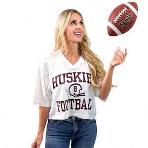 May include: A white short-sleeved mesh jersey featuring the maroon text HUSKIES FOOTBALL and a graphic of a football helmet with the number 8. A brown and white Franklin 100 football is positioned in the air above the jersey.