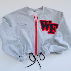 May include: A grey cropped sweatshirt with a red zip and black drawstrings. The sweatshirt features the embroidered word "Blackhawks" and a patch with the letters "WF" in black with a red border. The patch has three gold stars.