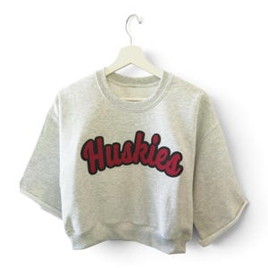 May include: A light gray heathered short-sleeve sweatshirt featuring the word Huskies in a bold, dark red script font with a thick black outline. The garment has a crew neckline, dropped shoulders, and wide, cuffed sleeves, hanging on a white hanger.