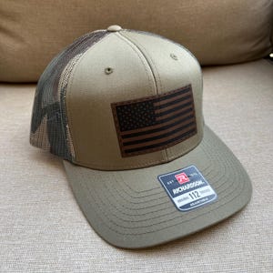 May include: A khaki-colored trucker hat with a brown leather patch featuring a distressed American flag. The hat has a mesh back and a curved brim. The Richardson 112 hat is adjustable.
