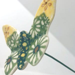 May include: A ceramic butterfly garden stake with a yellow and green floral design. The butterfly has white daisy-like flowers with brown centers and yellow accents. The wings are attached to a green wire.