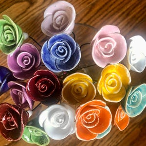 May include: A bouquet of 14 ceramic roses in various colors, including green, purple, blue, red, yellow, orange, white, and pink. The roses are arranged in a vase and are all facing the camera.
