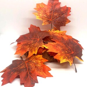 May include: A collection of artificial maple leaves in shades of red, orange, and yellow. The leaves have detailed veining and are arranged on a small wooden base, evoking an autumnal theme. Perfect for fall decor.