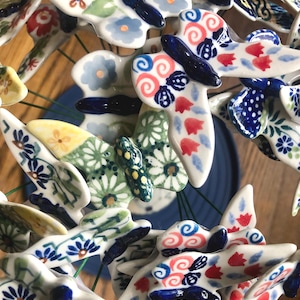 May include: A collection of ceramic butterfly ornaments in various patterns and colors. The butterflies feature designs with blue, red, and green floral and abstract motifs. The wings are white with detailed patterns, and the bodies are dark blue.