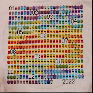 Può includere: Un motivo a punto croce colorato di case con i numeri da 1 a 12 e l'anno 2022. Le case sono disposte in file, ogni fila rappresenta un mese diverso dell'anno. Le case sono ricamate in una varietà di colori, tra cui rosso, arancione, giallo, verde, blu e viola.