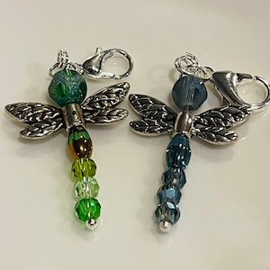May include: Two dragonfly charms with silver-tone wings and clasps. One dragonfly features a green bead head and a body of green and yellow beads. The other has a blue bead head and a body of blue beads. These charms are suitable for jewelry making.