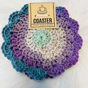 May include: A handmade, crocheted coaster in shades of teal, purple, and cream. The coaster has a scalloped edge and a tag that reads "COASTER" and "100% HANDMADE" with a coffee cup graphic.