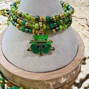 May include: Multi-strand bracelet with green and gold beads and a charm of a cartoon leprechaun wearing sunglasses and a hat.
