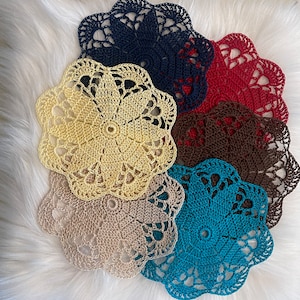 May include: A set of six crocheted doilies in various colors, including beige, cream, blue, brown, red, and navy blue. The doilies are arranged in a circle on a white fluffy surface.