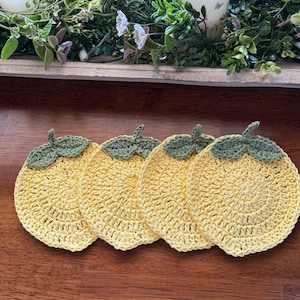 May include: Four handmade, yellow crochet coasters with green stems, arranged on a wooden surface. The coasters are round and have a textured, open weave design. The background includes greenery and candles.