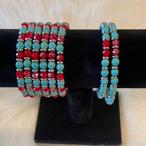 May include: Two beaded bracelets, one stacked on top of the other. The top bracelet is a stack of six bracelets, each with alternating turquoise and red beads. The bottom bracelet is a single bracelet with alternating turquoise and red beads.