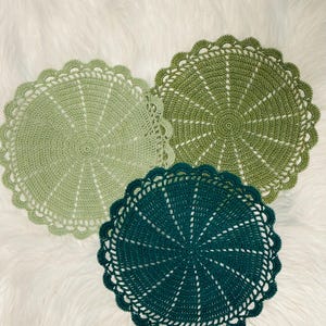 May include: Three handmade crochet doilies in shades of green and teal. Each doily features a circular design with a scalloped edge. The doilies are arranged on a white, fluffy surface.