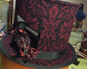 May include: A black top hat with a red and black damask pattern. The hat has a black ribbon and a red and black cross-shaped embellishment.