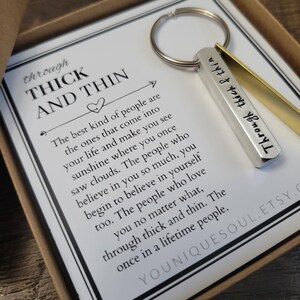 May include: A silver and gold keychain with the words "Through thick & thin" engraved on it. The keychain is in a box with a white card that reads "Through thick and thin. The best kind of people are the ones that come into your life and make you see sunshine where you once saw clouds. The people who believe in you so much, you begin to believe in yourself too. The people who love you no matter what, through thick and thin. The once in a lifetime people."