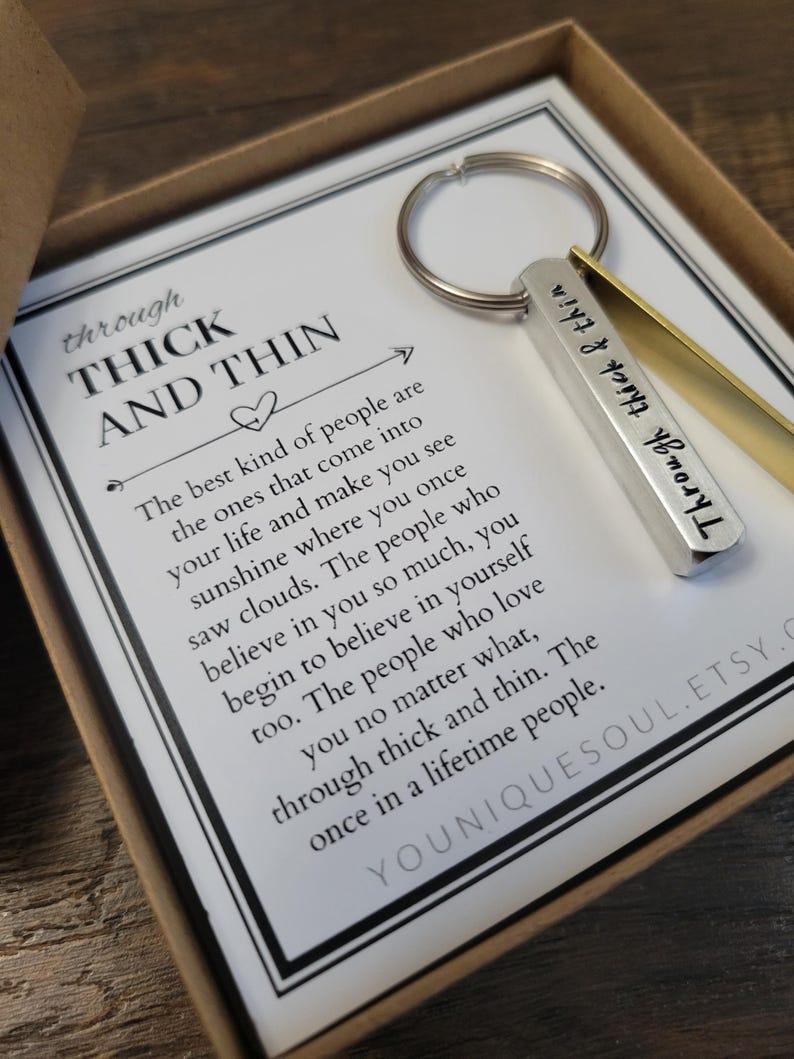 May include: A silver and gold keychain with the words "Through thick & thin" engraved on it. The keychain is in a box with a white card that reads "Through thick and thin. The best kind of people are the ones that come into your life and make you see sunshine where you once saw clouds. The people who believe in you so much, you begin to believe in yourself too. The people who love you no matter what, through thick and thin. The once in a lifetime people."