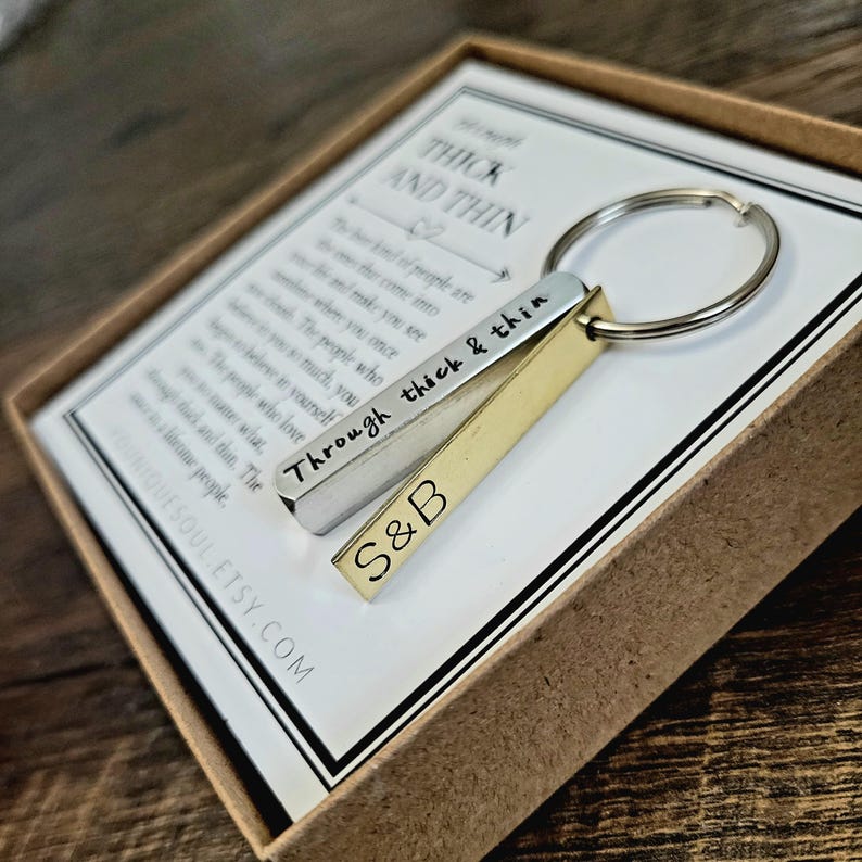 May include: A silver and gold keychain with the words "Through thick & thin" and "S&B" engraved on it. The keychain is in a small box with a quote about friendship.