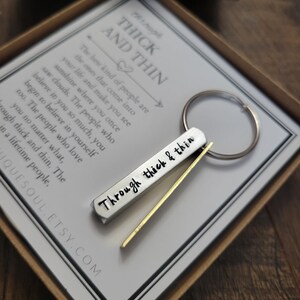 May include: A silver keychain with a gold bar attached. The keychain has the words "Through thick & thin" engraved on it. The keychain is in a box with a card that reads "The best kind of people are the ones that come into your life and make you see sunshine where you once saw clouds. The people who believe in you so much, you begin to believe in yourself too. The people who love you no matter what, through thick and thin. The lifetime people."