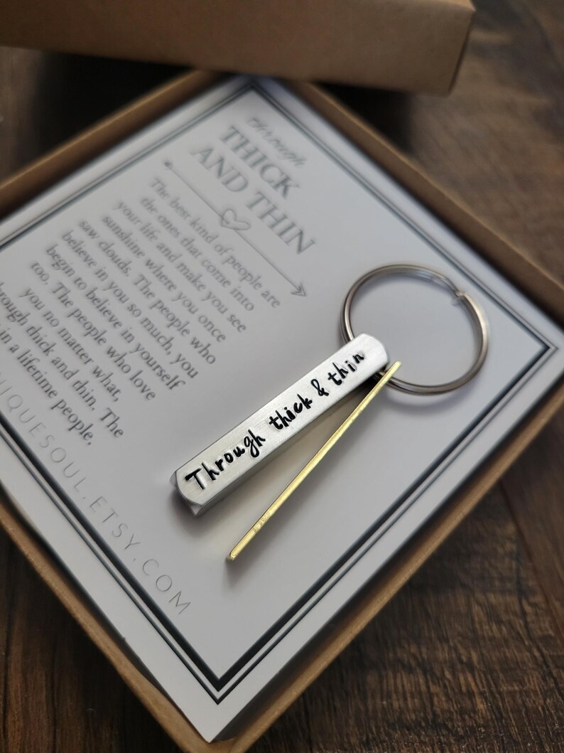 May include: A silver keychain with a gold bar attached. The keychain has the words "Through thick & thin" engraved on it. The keychain is in a box with a card that reads "The best kind of people are the ones that come into your life and make you see sunshine where you once saw clouds. The people who believe in you so much, you begin to believe in yourself too. The people who love you no matter what, through thick and thin. The lifetime people."