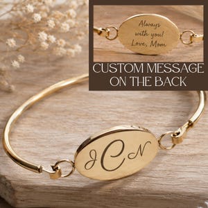 Puede incluir: Un brazalete dorado con una placa central ovalada grabada con las iniciales cursivas JCN. Un recuadro muestra el reverso de la placa con el texto grabado Always with you! Love, Mom, y el texto CUSTOM MESSAGE ON THE BACK en la imagen.