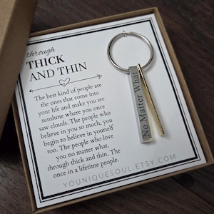 May include: A silver-toned metal keyring with a rectangular bar pendant engraved with the text No Matter What, paired with a thin gold-toned bar, attached to a circular ring, presented on a card with the text through THICK AND THIN.