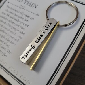 May include: A silver and gold keychain with the words "Through thick & thin" engraved on it. The keychain is attached to a silver key ring. The keychain is in a box with a white card that says "Through thick and thin" and a quote about the best kind of people.