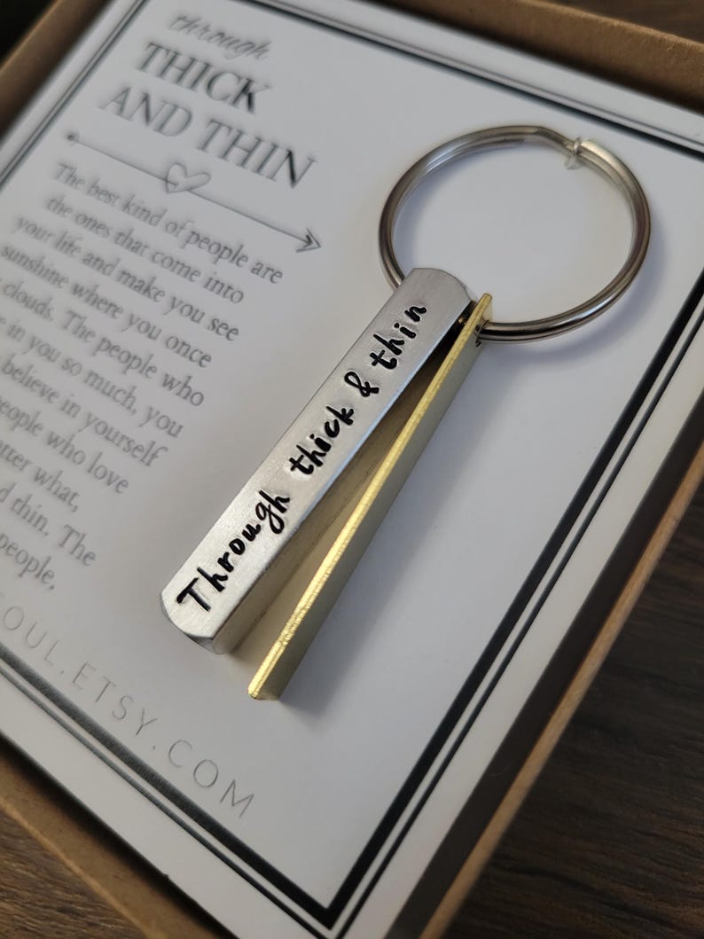 May include: A silver and gold keychain with the words "Through thick & thin" engraved on it. The keychain is attached to a silver key ring. The keychain is in a box with a white card that says "Through thick and thin" and a quote about the best kind of people.