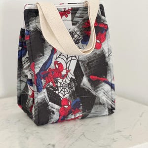 May include: A rectangular fabric lunch bag featuring a repeating pattern of the superhero Spider-Man in red and blue, set against a black and white cityscape background with spiderweb motifs. The bag has two beige canvas handles at the top.