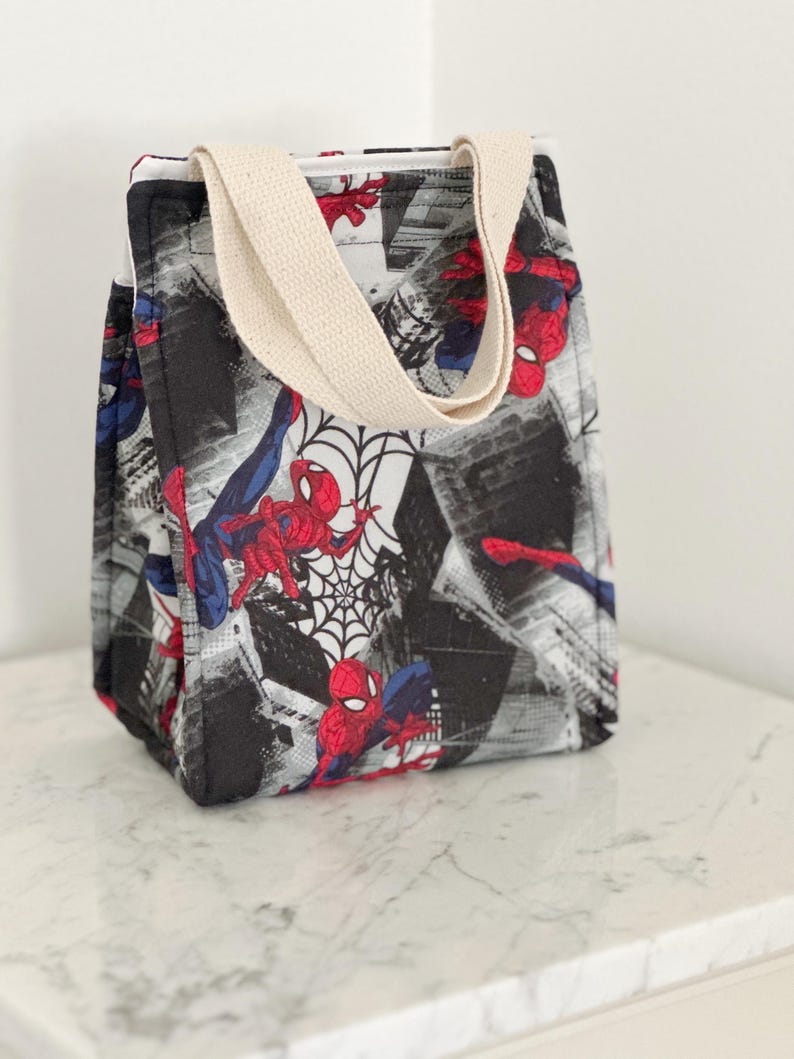 May include: A rectangular fabric lunch bag featuring a repeating pattern of the superhero Spider-Man in red and blue, set against a black and white cityscape background with spiderweb motifs. The bag has two beige canvas handles at the top.