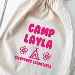 May include: A beige drawstring bag with the text "CAMP LAYLA" in bright pink, along with a tent and flower graphics. The words "SLEEPOVER ESSENTIALS" are also printed in pink. The bag has a drawstring closure.