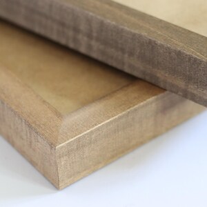 May include: Two wooden picture frames, one on top of the other. The top frame is a darker brown wood with a smooth finish. The bottom frame is a lighter brown wood with a more textured finish.