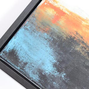 May include: Abstract art piece with a black frame. The artwork features a blend of colors, including orange, blue, and gray, creating a textured, layered effect. The painting is mounted in a dark wooden frame, adding a modern touch.
